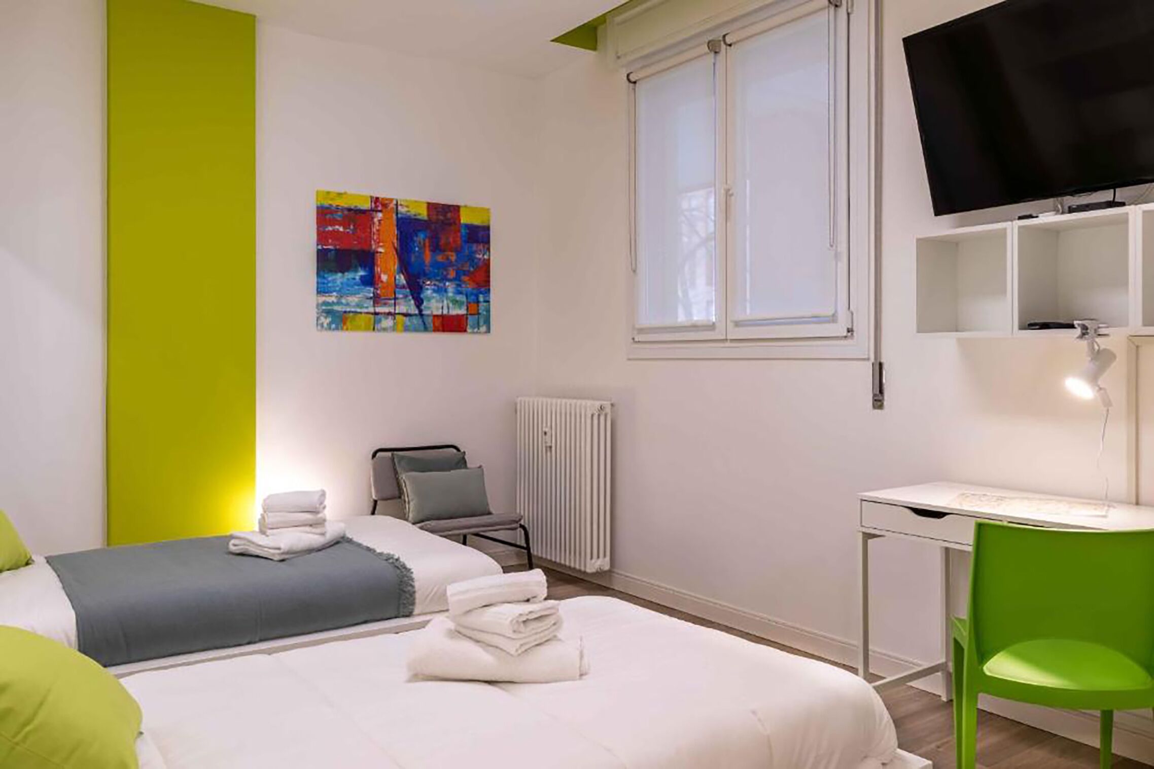 Une chambre d’hôtel avec deux lits, un bureau, une chaise, un radiateur, une télévision et un tableau coloré au mur.