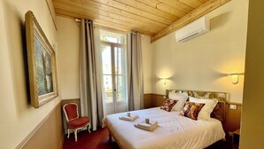 Una habitación de hotel con cama, sillón, un cuadro, ventana con cortinas y equipo de aire acondicionado.