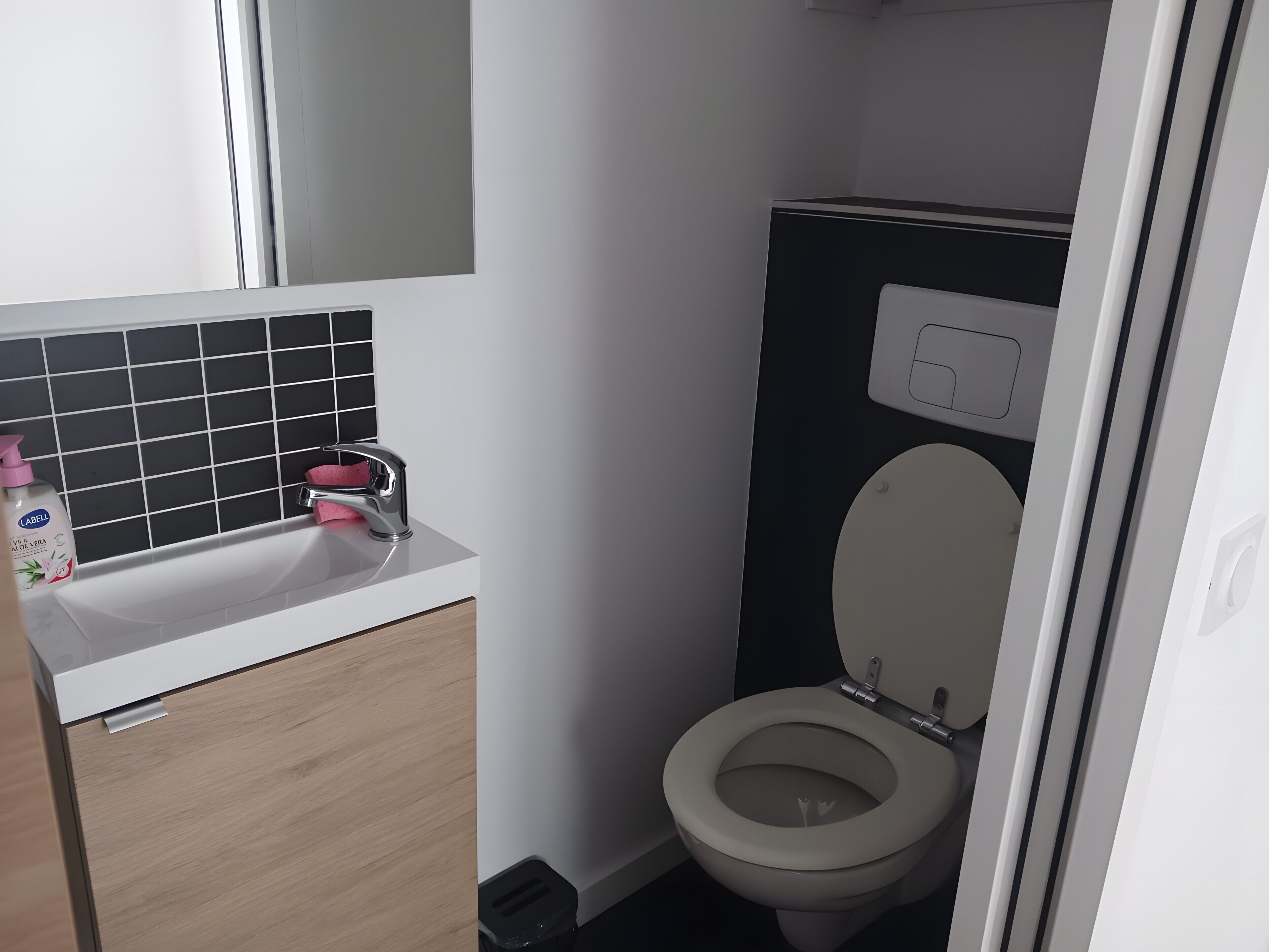 Ein Badezimmer mit weißem Waschbecken, einem Spiegel, einer schwarzen Fliesenwand, einer Toilette und einem Mülleimer.