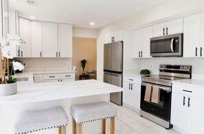 Eine moderne Küche mit weißen Schränken, einer zentralen Kochinsel mit Hockern, einem Kühlschrank, einer Mikrowelle und einem Backofen.