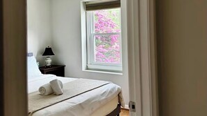 A bedroom with a bed, a nightstand, a lamp, and a window with pink flowers outside.