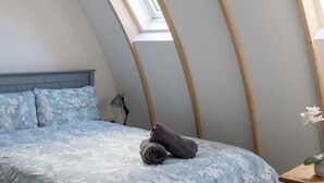 Ein Schlafzimmer mit schrägverdachter Decke, einem Bett mit floralem Überwurf, einem zusammengerollten Handtuch, einer Nachttischlampe und einem kleinen Tisch mit einer Blume.