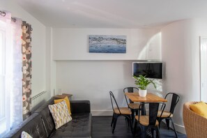 Ein Wohnzimmer mit einer Couch, einem Esstisch mit Stühlen, einem an der Wand montierten Fernseher und einem Gemälde darüber.