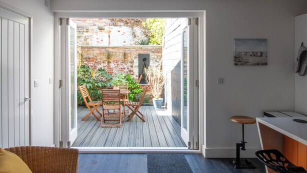Eine offene Schiebetür führt zu einer Terrasse mit einem Holztisch und Stühlen, einem kleinen Hocker und einem Blumentopf.