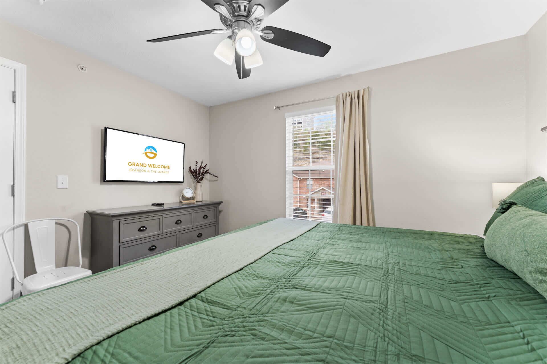 Une chambre à coucher avec un lit, un ventilateur de plafond, une commode, un téléviseur et une fenêtre avec des rideaux.