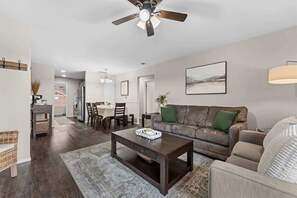 Ein modernes Wohnzimmer mit einem dunkelholzigen Couchtisch, einem grauen Sofa mit grünen Kissen, einem Essbereich mit einem weißen Tisch und einem Deckenventilator.