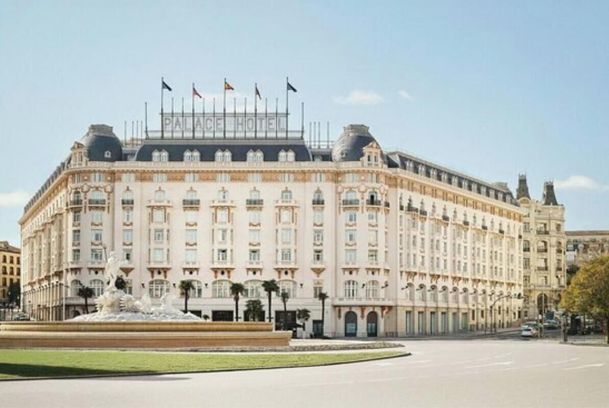 A large, historic building with a central fountain, multiple flags, and a symmetrical facade.