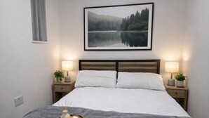 A neatly made bed with a grey blanket, two bedside tables with lamps, a framed picture above the bed, and a tray with candles on the bed.