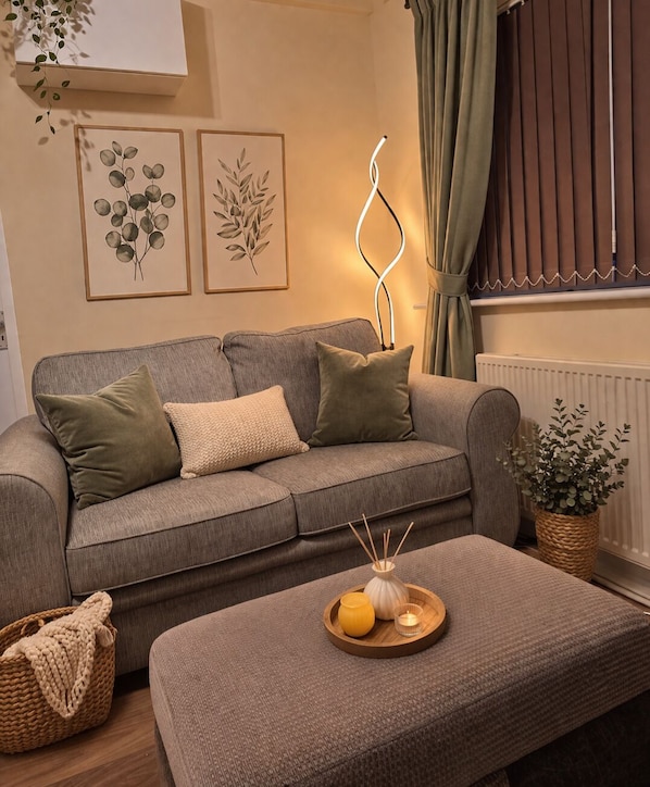 A living room with a grey sofa, a wooden side table with a candle and a vase, and framed botanical prints on the wall.