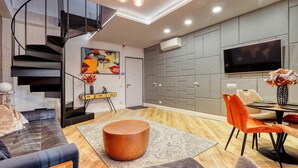 Ein modernes Wohnzimmer mit Treppe, Flachbildfernseher, Essbereich mit orangefarbenen Stühlen und einem großen Gemälde an der Wand.