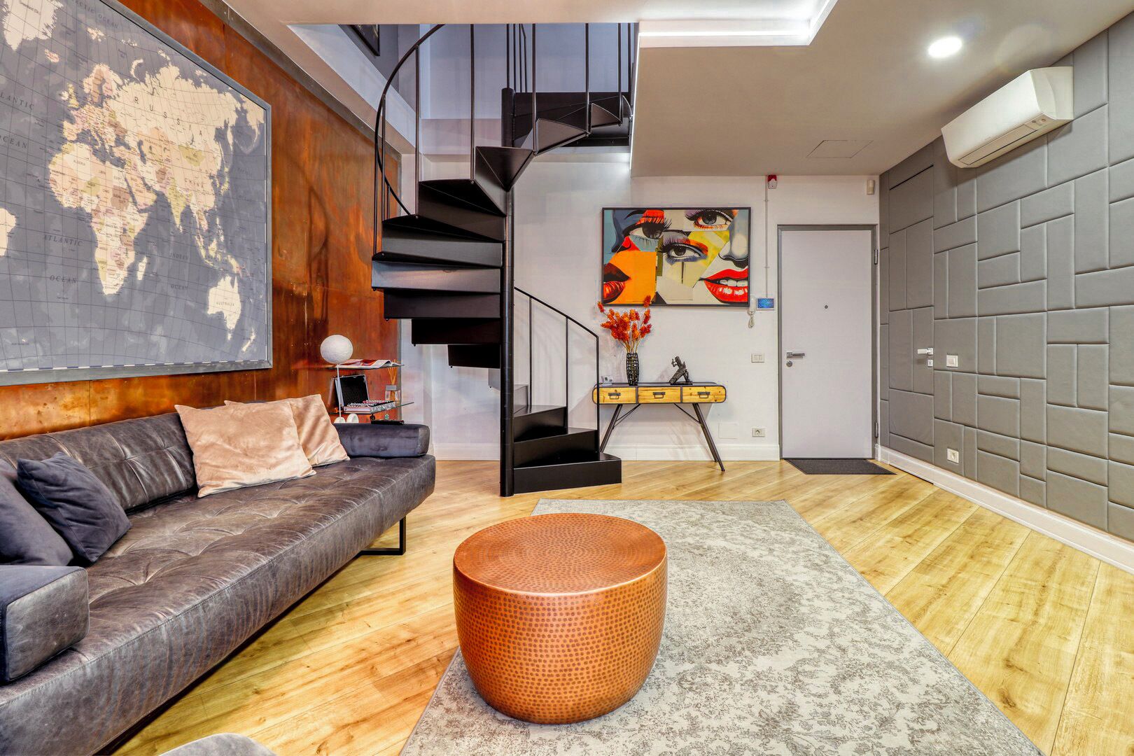 A modern living room with a leather sofa, a round wicker ottoman, a world map on the wall, a painting, and a staircase.