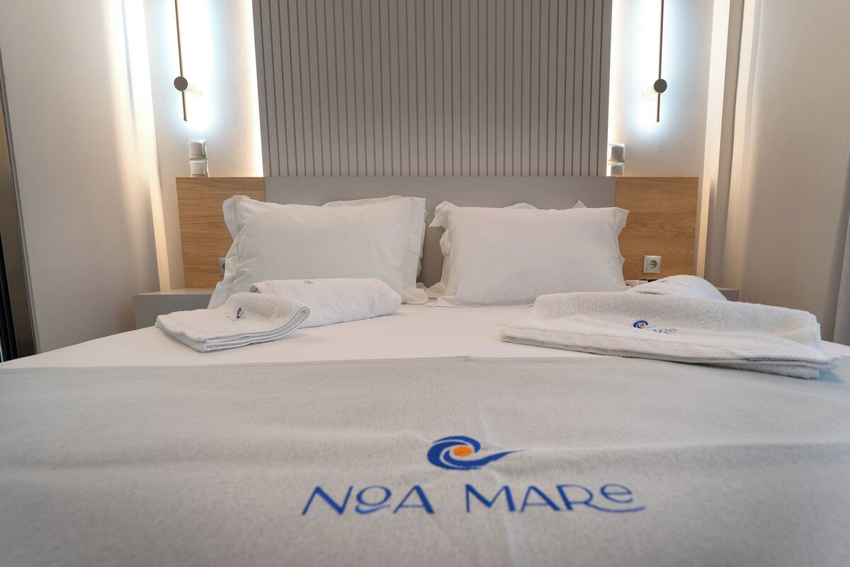 Un lit bien fait, avec des serviettes pliées et une couvre-lit à l’effigie de la marque, une tête de lit en bois et un luminaire fixé au mur.