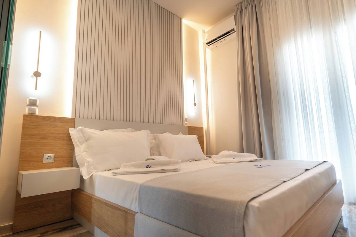 Une chambre d’hôtel avec un grand lit, une tête de lit en bois, une table de chevet, une commode et une fenêtre avec des rideaux.