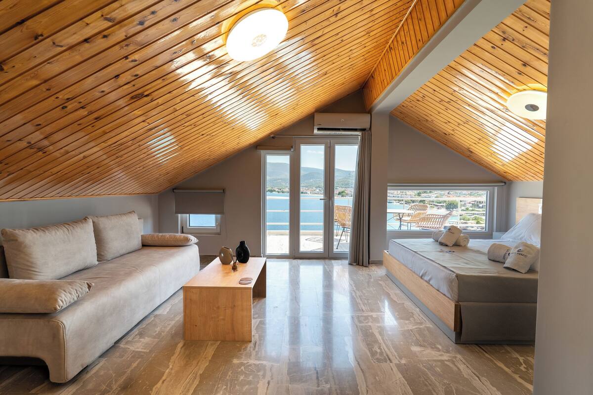 Une chambre spacieuse aménagée en mezzanine, avec un plafond en bois, un grand lit, un canapé et une vue sur la mer.