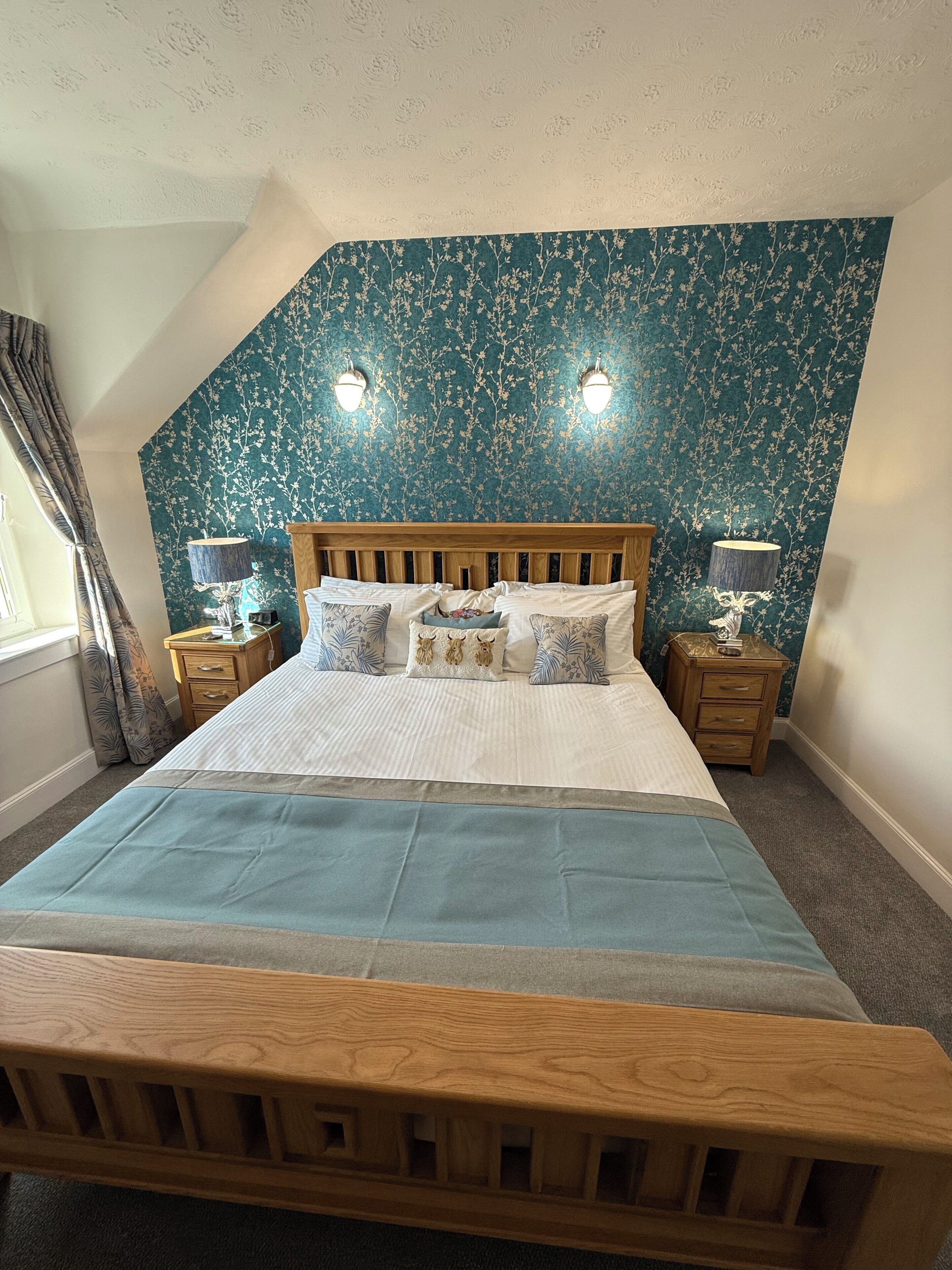 A bedroom with a wooden bed, floral wallpaper, two bedside lamps, and a window with curtains.
