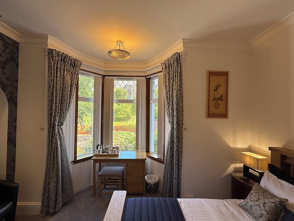 A bedroom with a bed, a desk, and a chair. There is a window with curtains, a lamp, and a framed picture on the wall.