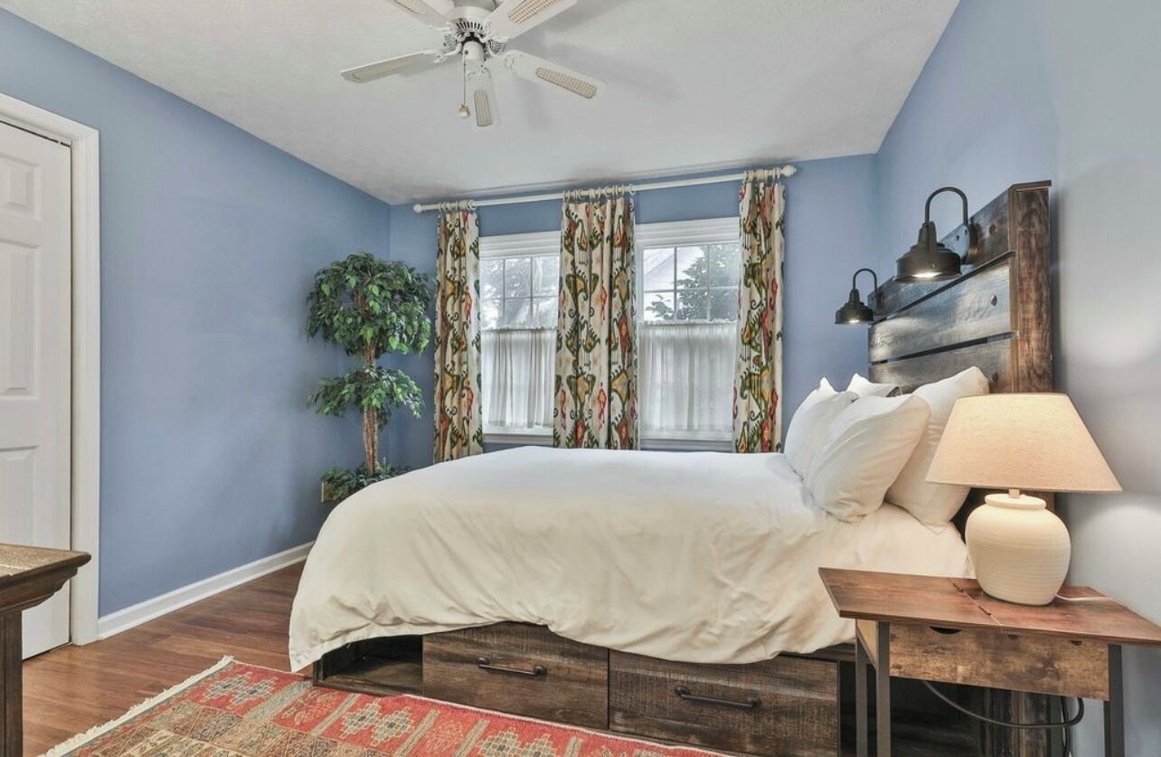 A bedroom with a bed, a nightstand, a lamp, a ceiling fan, and a window with curtains.
