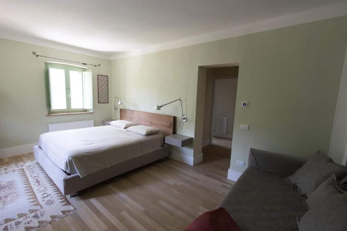 Un dormitorio con cama, mesitas de noche, ventana, puerta y sofá.