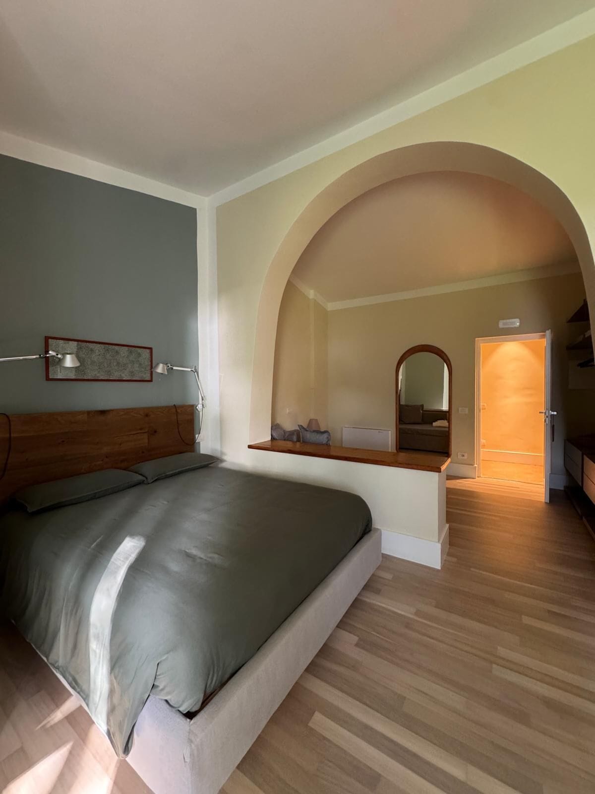 Een slaapkamer met een bed, een houten hoofdeinde, een nachtkastje, een spiegel en een gewelfde deuropening naar een andere kamer.