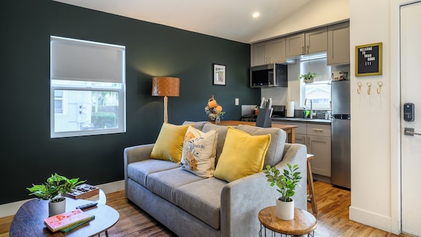 Ein modernes Wohnzimmer mit grauem Sofa, einem kleinen runden Couchtisch und einer Küchenzeile mit grauen Schränken und einer schwarzen Mikrowelle.