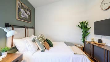 A neatly made bed with pillows, a bedside table with a lamp, a wall-mounted clock, and a television on a wooden stand.