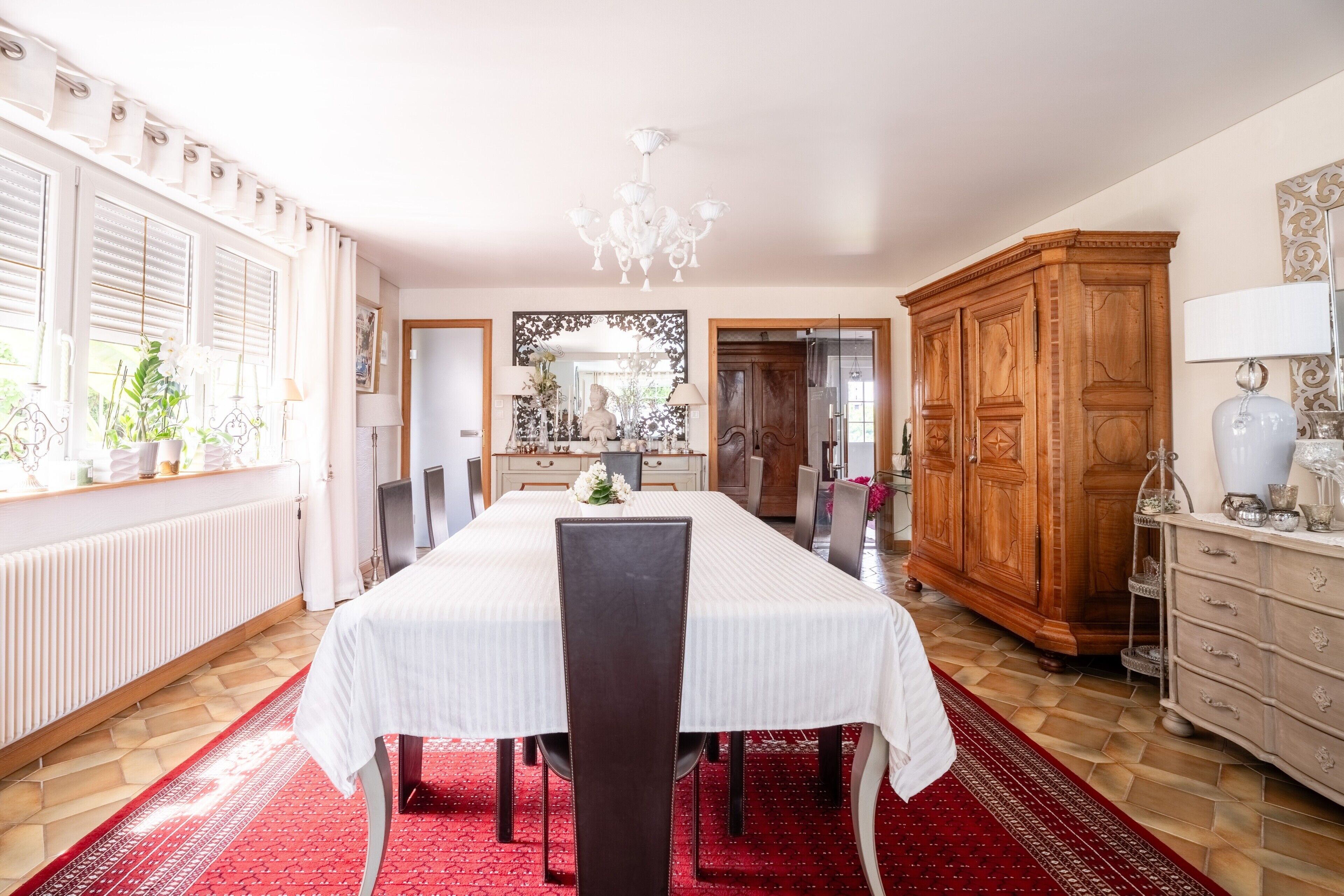 Un espace repas comprenant une longue table, des chaises, un lustre, un buffet en bois et un radiateur.