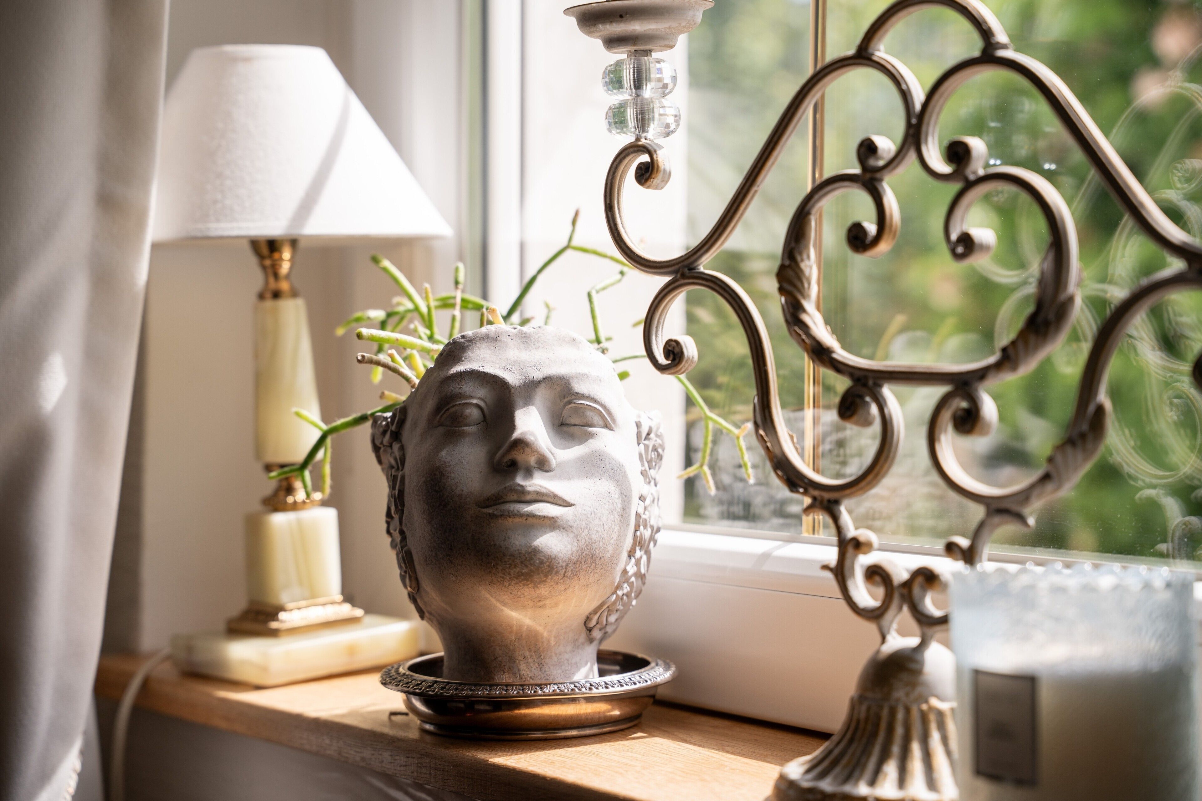 A window with a decorative metal frame, a white lamp, and a vase with greenery on a wooden surface.