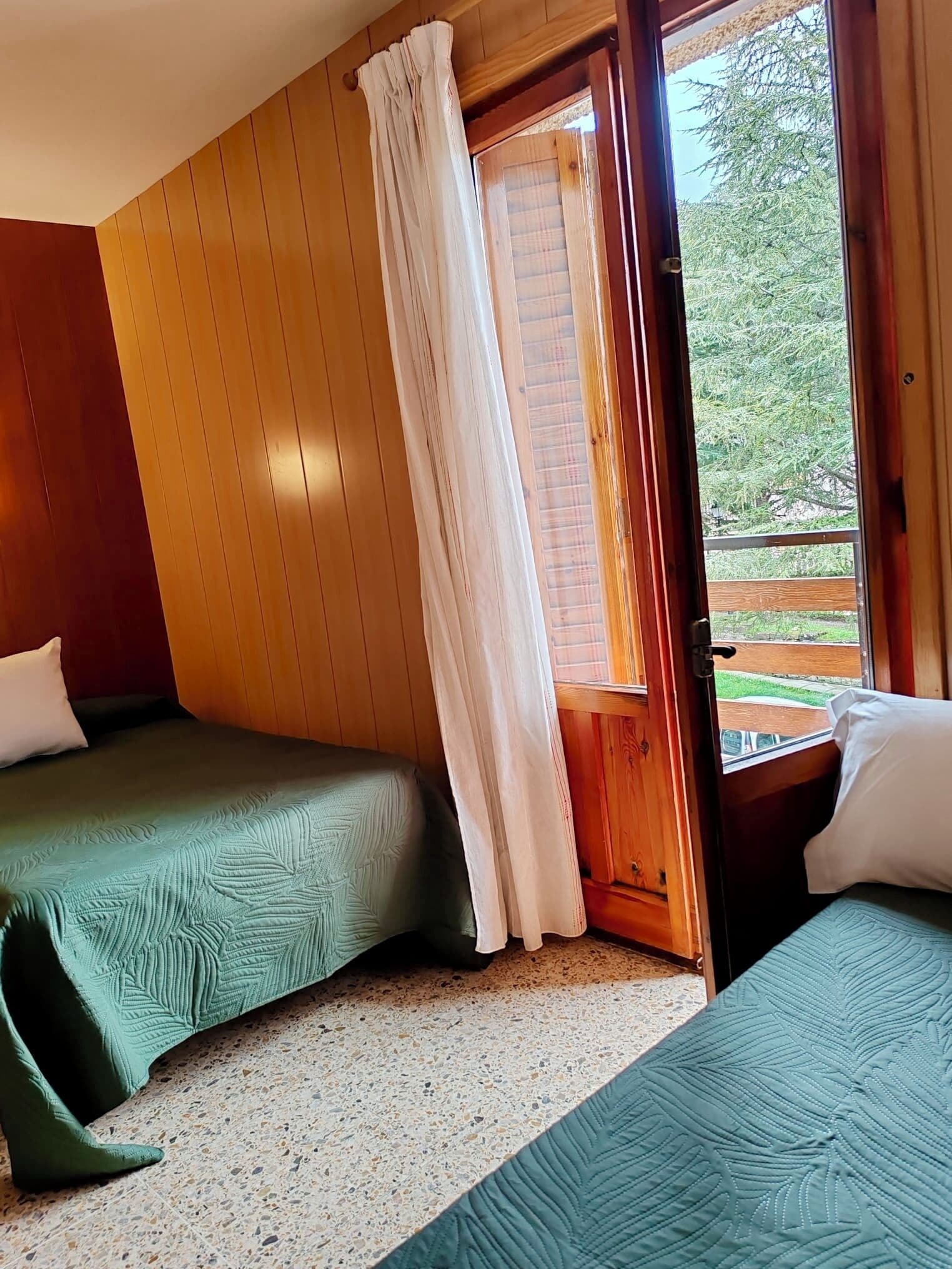 Una habitación con dos camas, paredes de madera, una ventana con cortinas y vistas al verdor exterior.
