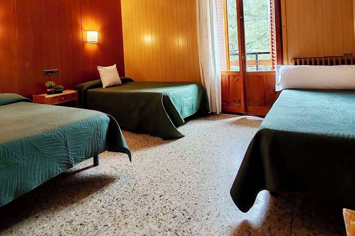 A room with wooden walls, two beds with green bedding, a small table with a lamp, and a window with curtains.