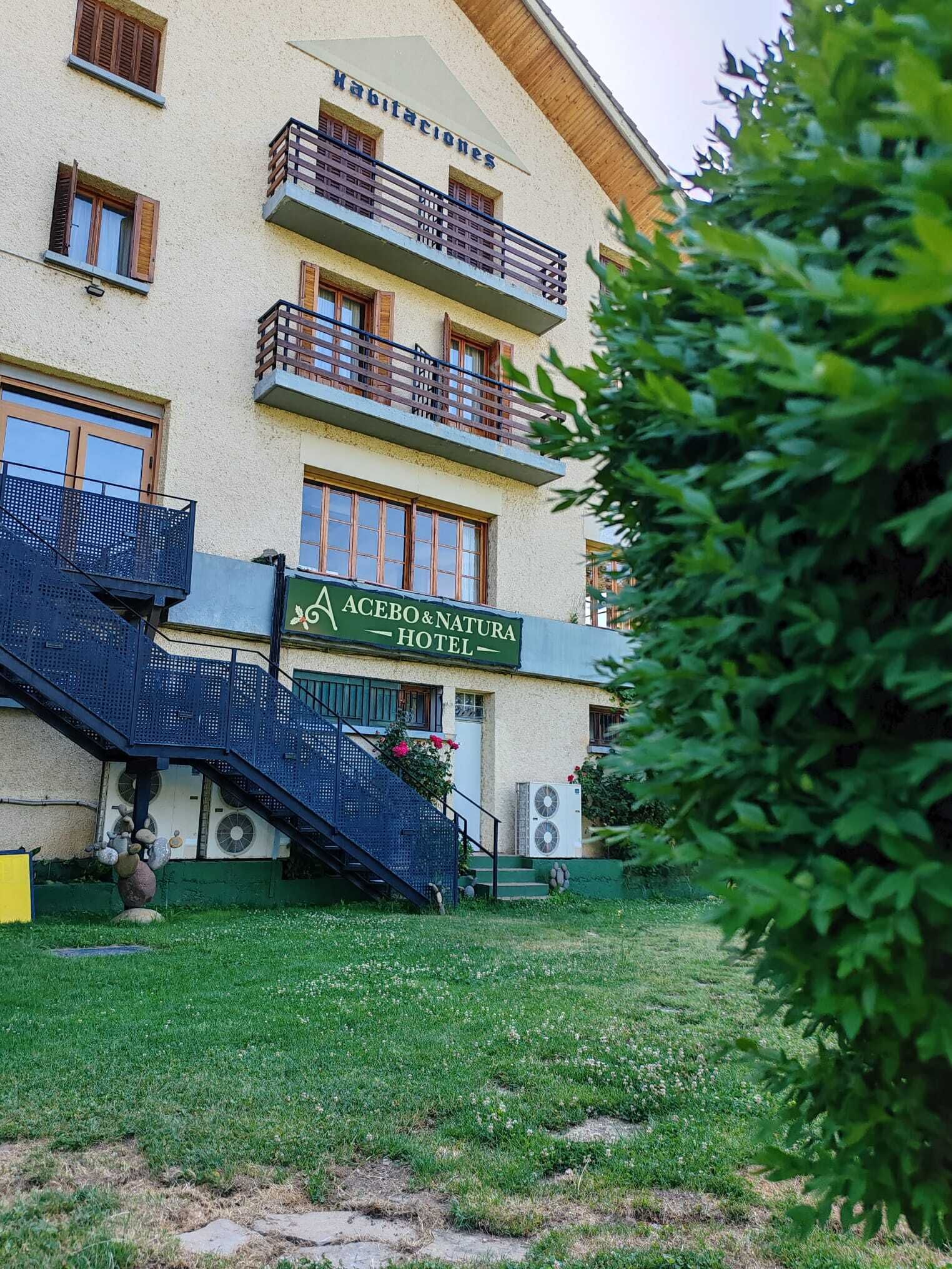 A hotel with a sign reading 'ACEBO & NATURA HOTEL,' a staircase leading to the entrance, and a green lawn in the foreground.