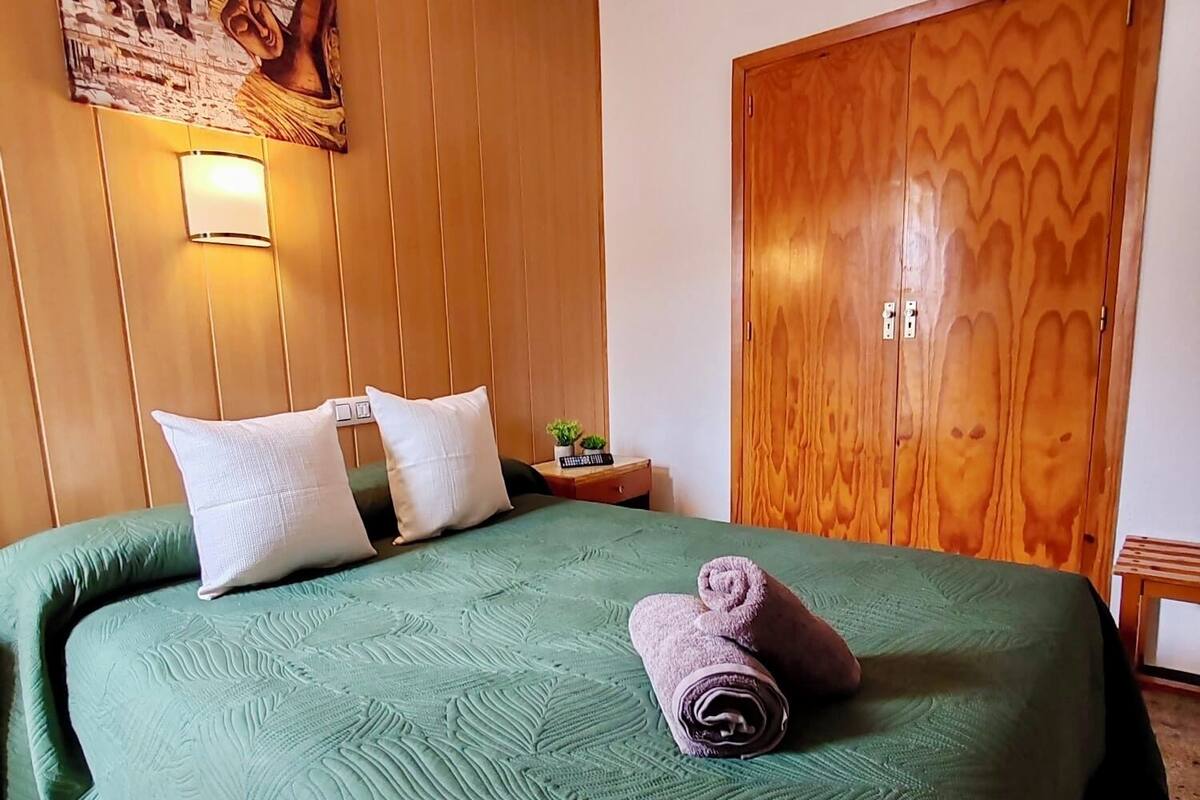 A hotel room with a green bed, pillows, a wall-mounted lamp, a wooden wardrobe, and a painting.