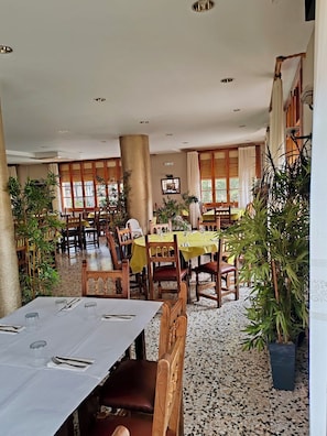 Un espace repas avec des tables préparées pour deux personnes, des chaises en bois et des plantes en pot.