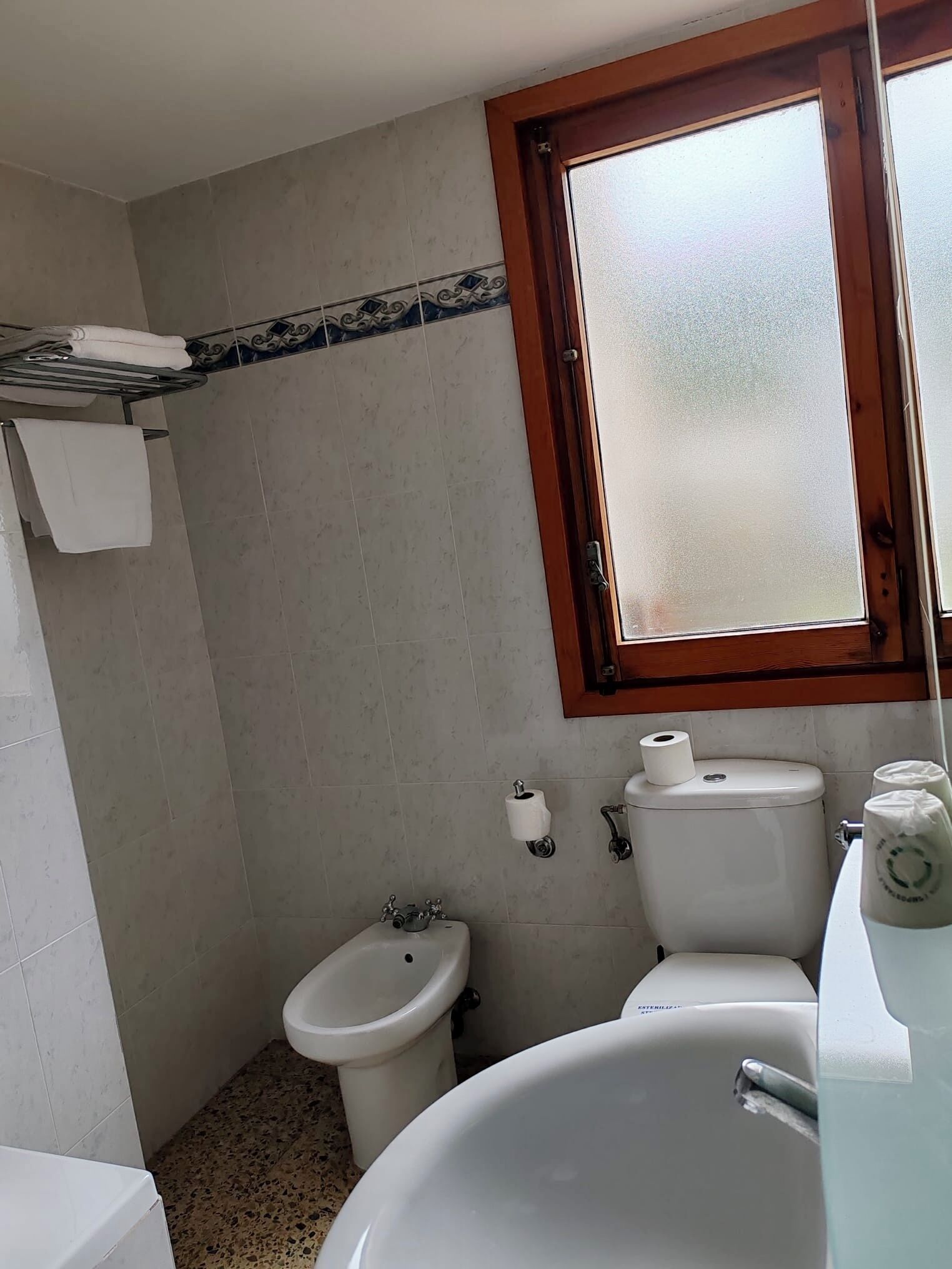 A compact bathroom with a toilet, bidet, and sink. A window with a wooden frame is above the sink.