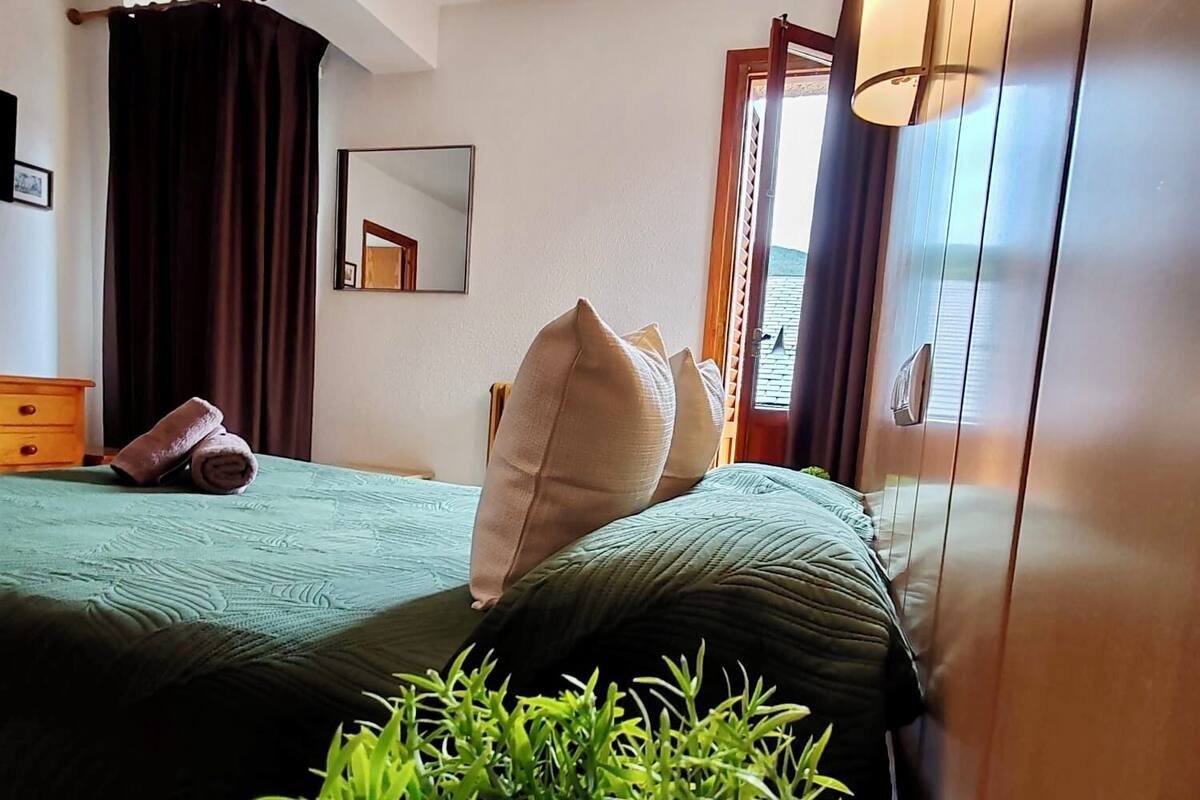 A hotel room with two beds, a wooden nightstand with a plant, a mirror, and a window with curtains.