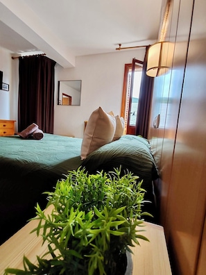 Une chambre d’hôtel avec deux lits, une table de chevet en bois ornée d’une plante, un miroir et une fenêtre avec des rideaux.