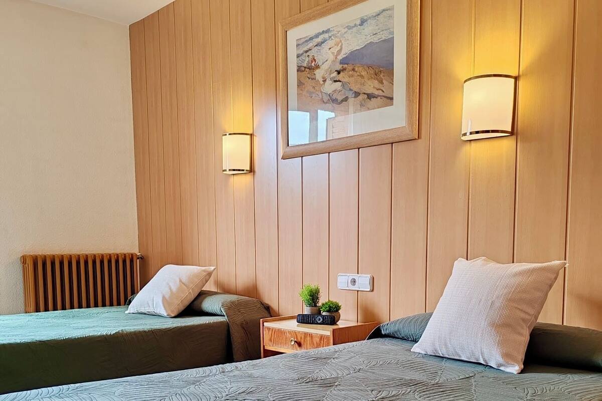 A hotel room with two beds, wooden panel walls, a painting, two wall-mounted lamps, a nightstand with a plant, and a radiator.