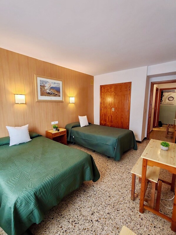 Una habitación con dos camas, una puerta de madera, una pequeña mesa con una planta y un cuadro enmarcado en la pared.