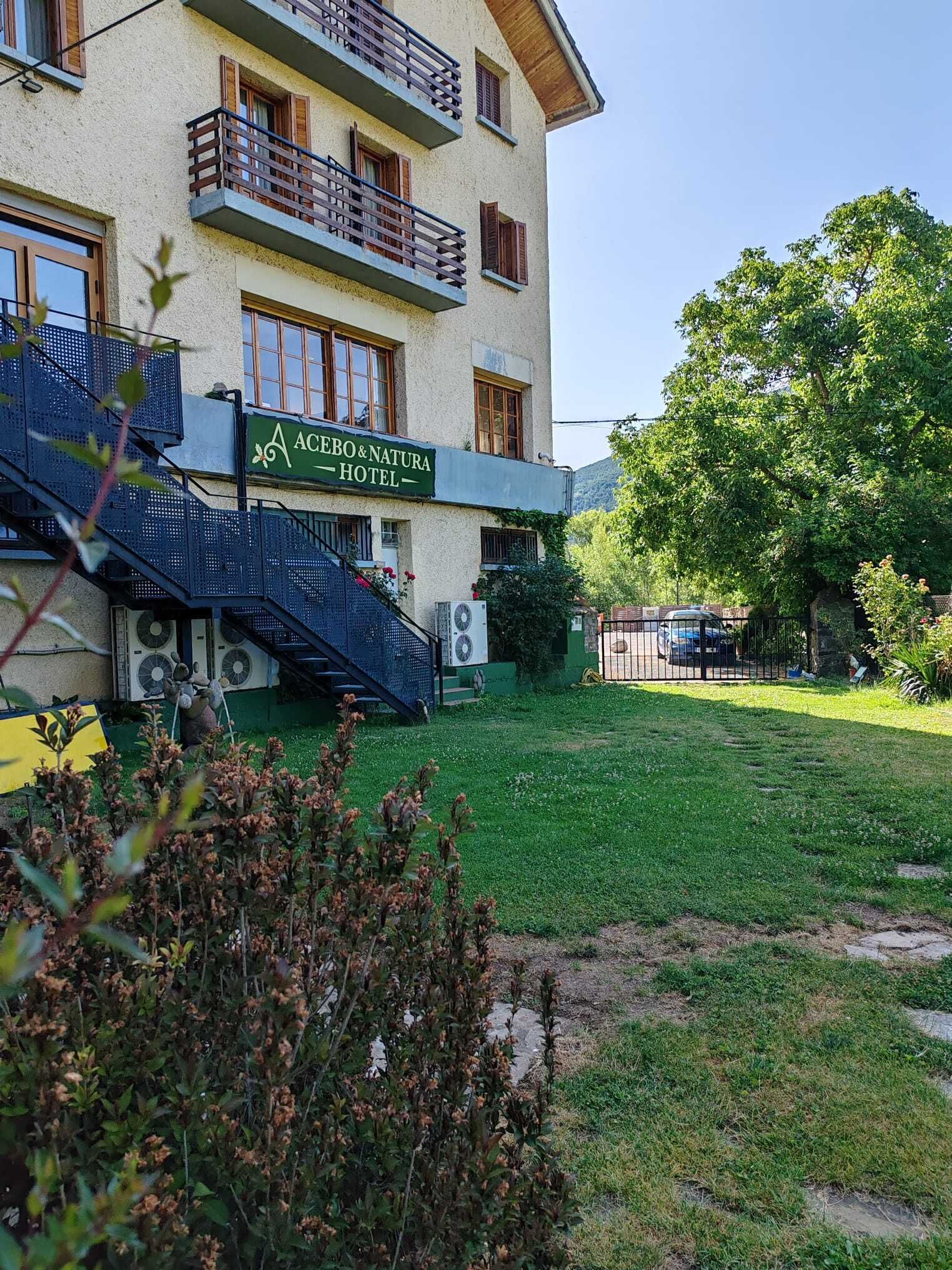 A hotel with a sign reading 'AEBEO & NATURA HOTEL,' a staircase, and a well-maintained lawn.