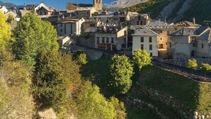 Un pueblo en una ladera con edificios de piedra, una torre de iglesia y un muro de piedra.