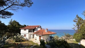 Una casa de dos pisos con techo de tejas rojas, rodeada de vegetación y vista al mar.