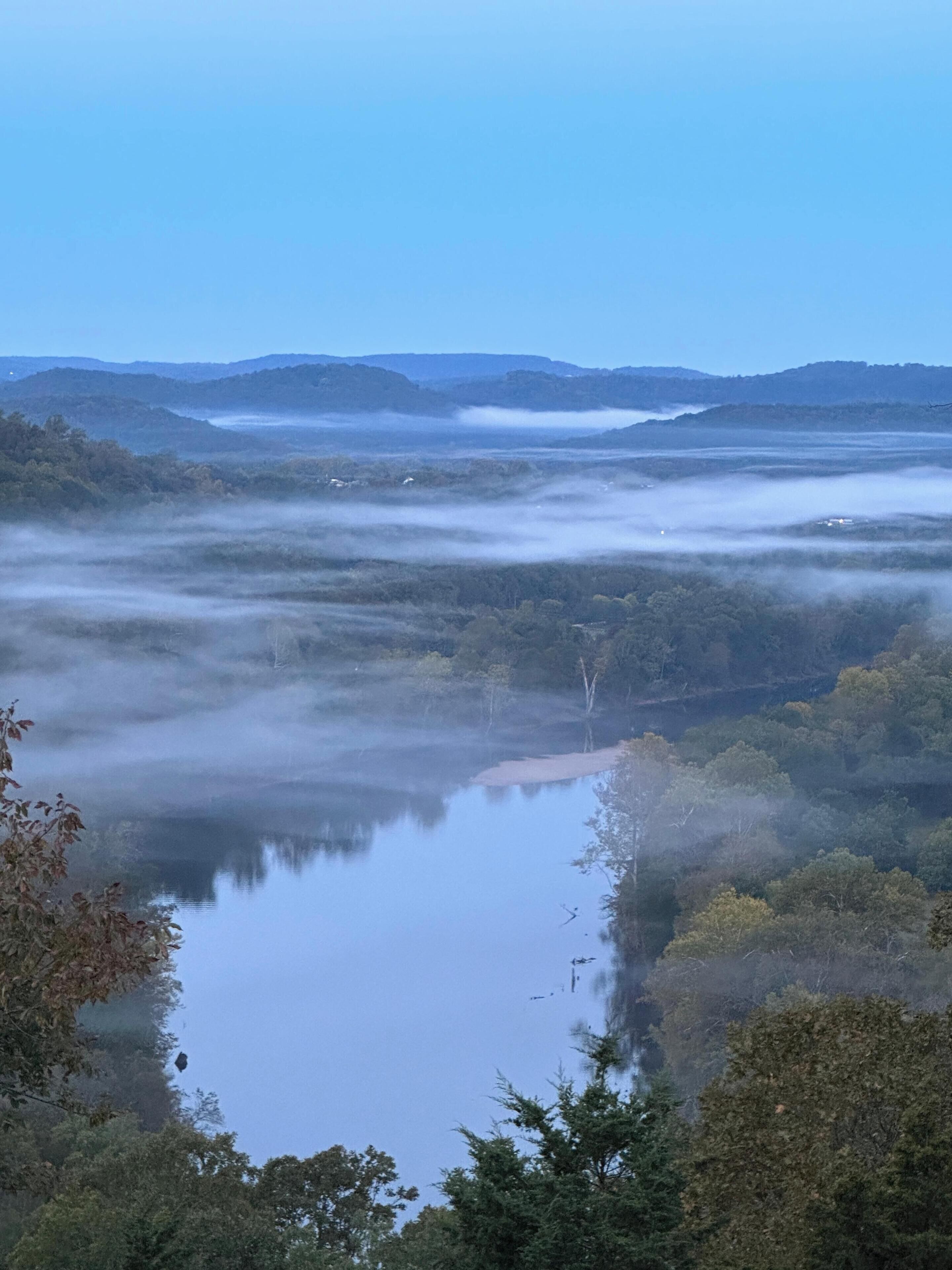 A misty landscape with a calm lake, surrounded by dense forest and distant hills.