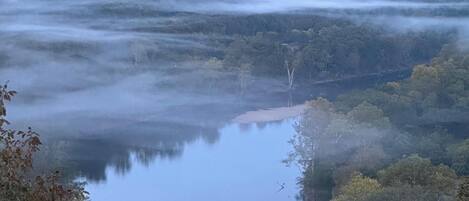 A misty landscape with a calm lake, surrounded by dense forest and distant hills.