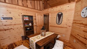 Interior de una cabaña de madera con una mesa, bancos, un farol, una estantería con libros y un reloj en la pared.