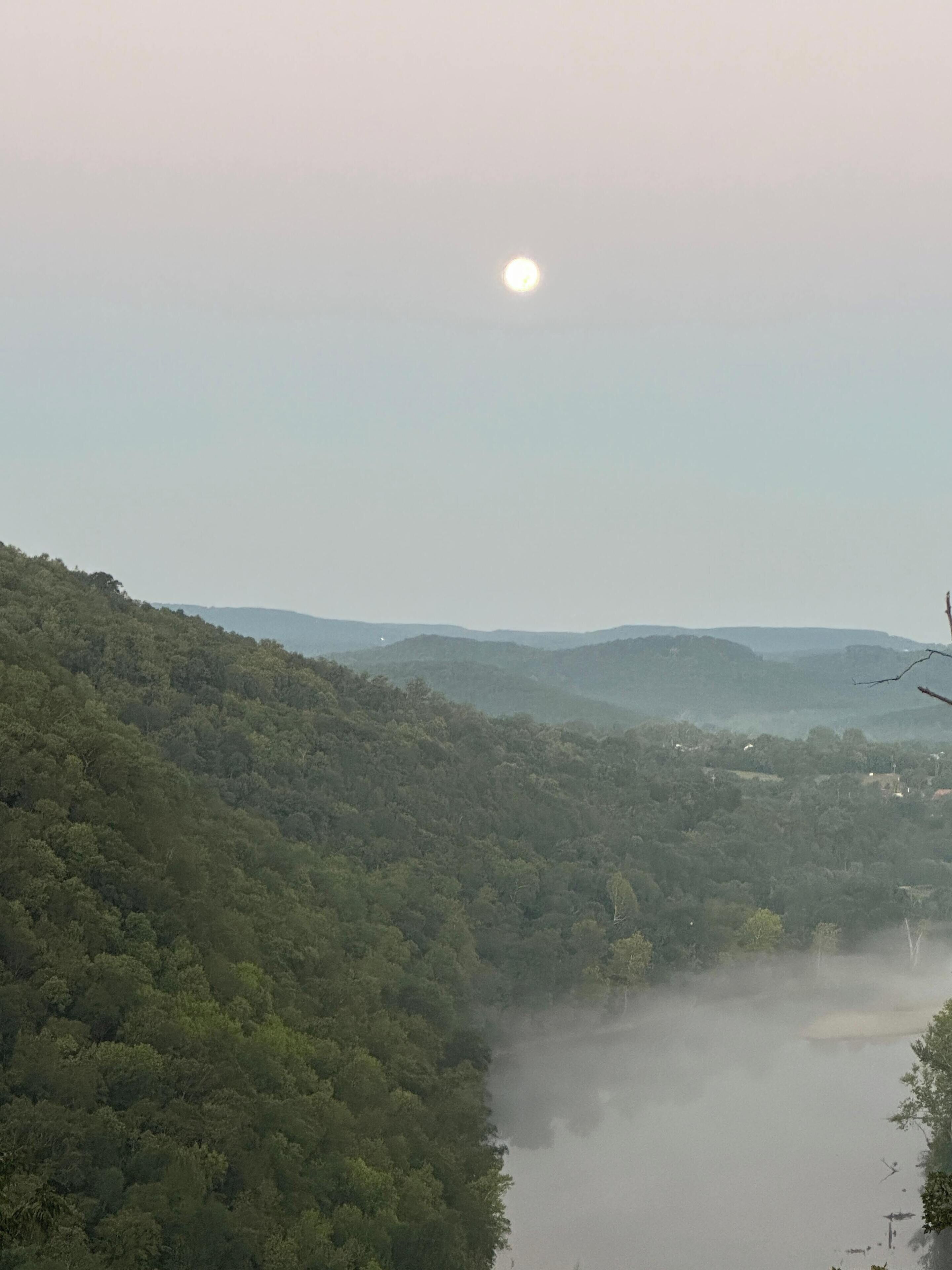 Un paysage montagneux avec une forêt dense, une rivière et une lune bien visible.