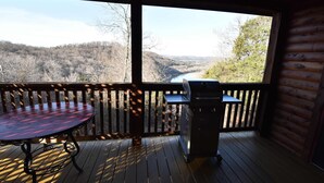 Una terraza de madera con una mesa y una parrilla, con vistas a un río y colinas boscosas.