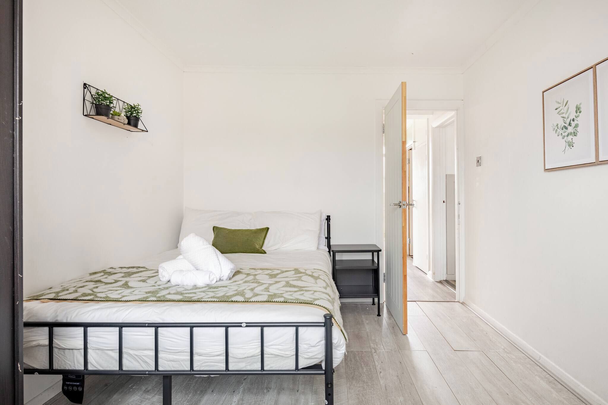 A neatly arranged bedroom with a bed, bedside table, and a small shelf with plants.