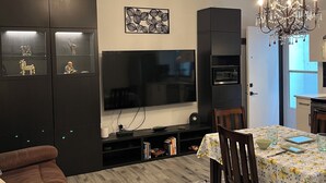 Ein Wohnzimmer mit einem braunen Sofa, einem Esstisch mit Stühlen, einer Kristallleuchter, einem Flachbildfernseher und einer schwarzen Unterhaltungsanlage mit Regalen.