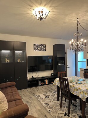 Un salon avec un canapé marron, une table à manger avec des chaises, un lustre, un téléviseur à écran plat et un meuble de rangement noir avec des étagères.