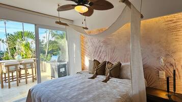Un dormitorio con cama, ventilador de techo, balcón con mesa y sillas, y vista a palmeras.