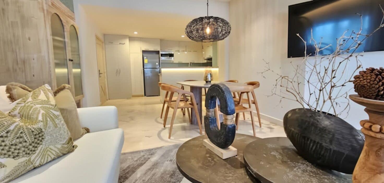 Een moderne woonkamer met een bank, een eettafel, stoelen, een opvallende sculptuur en een aan de muur bevestigde televisie.